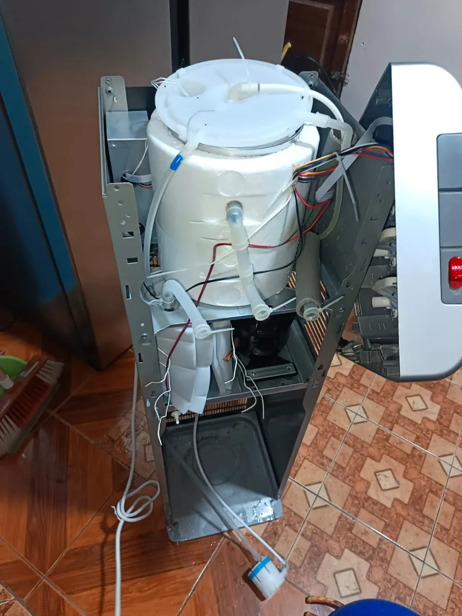 Dispensador de agua abierto por un lado, con un depósito blanco, tubos transparentes y cables a la vista
