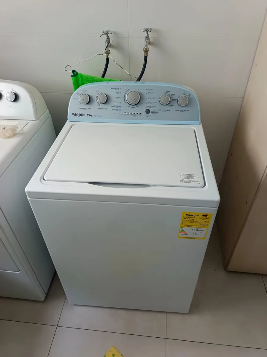 Lavadora de carga superior blanca instalada en un lavandero, con las llaves de agua detrás