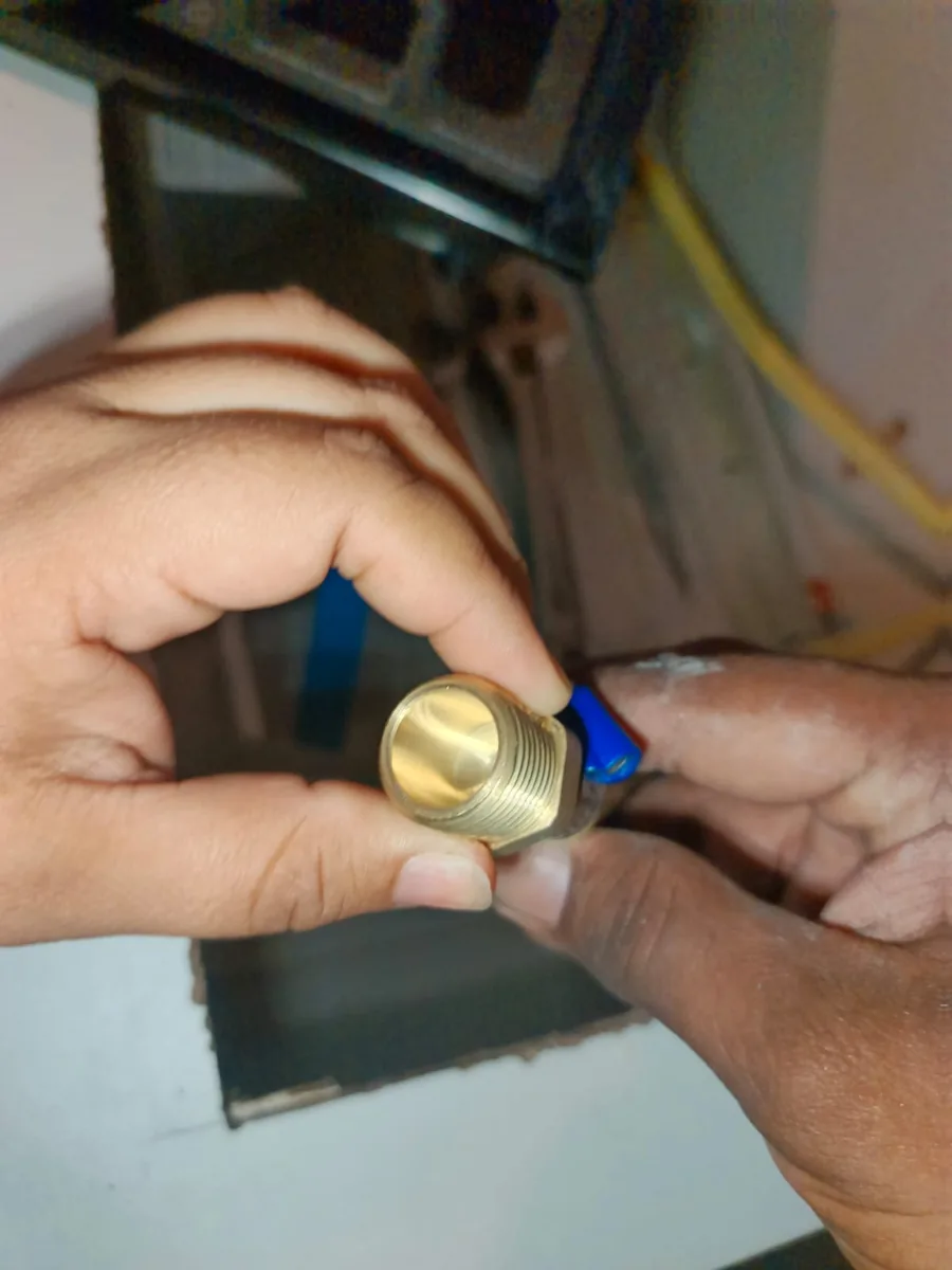 Manos sosteniendo una pieza de latón dorada con una palanca azul, frente a una manguera amarilla
